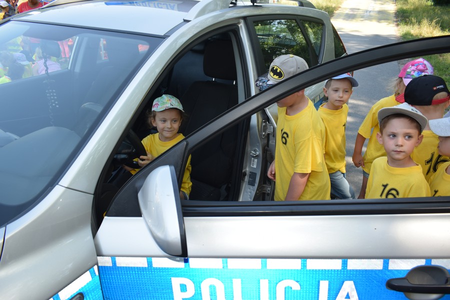 Grupa najmłodszych w żółtych t-shirtach obok samochodu osobowego z dużym niebieskim napisem na drzwiach „Policja”. Zadowolone dzieci zajmują kolejno miejsce za kierownicą pojazdu.