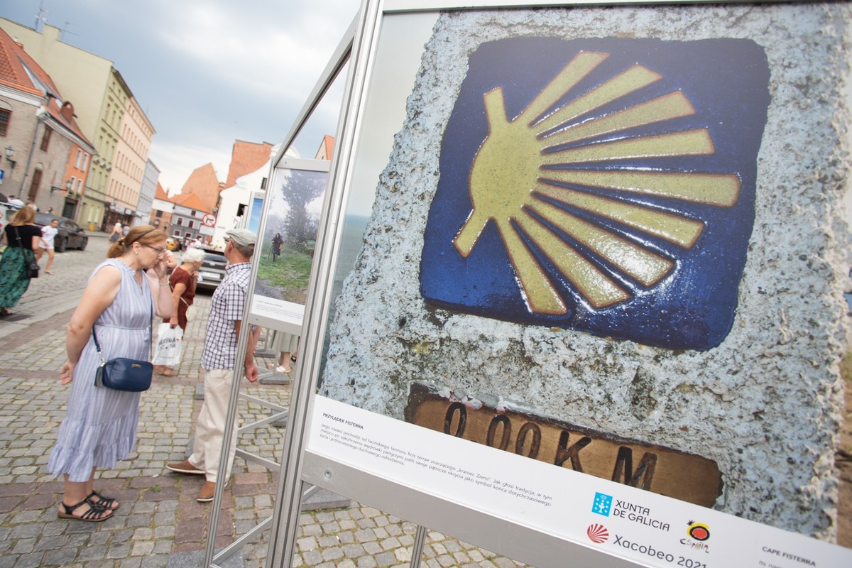 Na zdjęciu widać wystawę fotograficzną umieszczoną na ulicy. W centrum zdjęcia znajduje się zbliżenie na zdjęcie przedstawiające muszlę na szlaku Camino de Santiago. W tle można zauważyć kilka osób oglądających wystawę. Całość odbywa się w scenerii miejskiej, z brukowaną ulicą i zabytkowymi budynkami w tle.