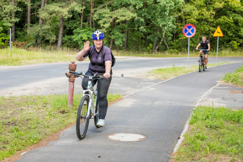 Dwójka rowerzystów jadących w pewnej odległości od siebie na asfaltowej ścieżce rowerowej przy ulicy w leśnej scenerii